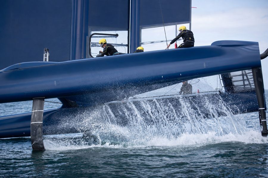 A foiling sailboat with its hull still in the water, spray over the bow